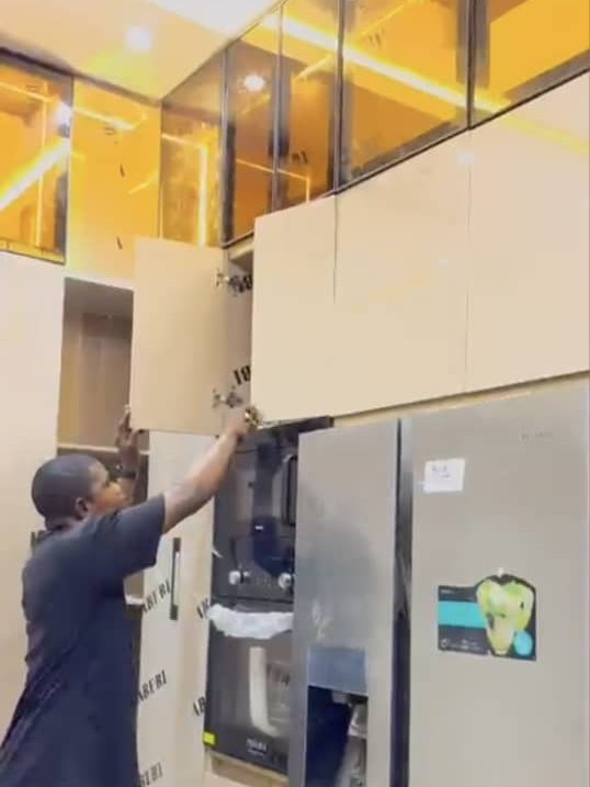A fitter hinging the door of a tall unit beside a fridge, under a lit gold glass upper run